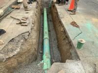 A turquoise sewer pipe is set inside a gravel-lined trench cut into the pavement of the road.