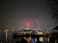 Fireworks in the distance behind a sailboat with holiday lights strung along its rigging.