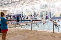 Minnie Howard's therapeutic pool featuring a group of community members engaged in an aquatics program, while a lifeguard stands on the left side facing them, another lifeguard walks between the two pools, and a third lifeguard sitting on his lifeguard chair