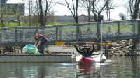 Woman on the deck while holding onto a kayak; another woman in a kayak on the launch with both arms up holding a paddle and smiling wide