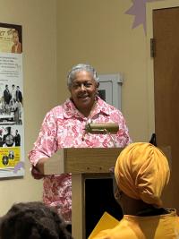 A person speaking at a podium in a room decorated with posters. One poster features historic photographs. People in the audience are visible, with one wearing a yellow head covering.
