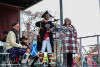 town crier on stage with a scroll surrounded by two people, one on the left sitting and one on the right standing