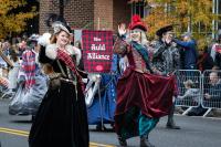 two women in elaborate dresses in hats walking in the parade holding a banner that says the Auld Alliance