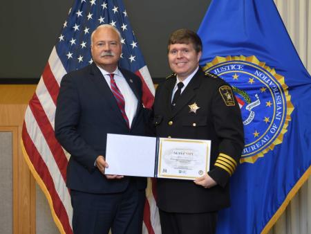 Man in business attire presenting certificate to Sheriff in formal dress uniform with U.S. flag and FBI flag in background