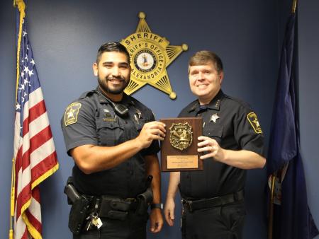 Deputy standing with sheriff, both in blue uniforms, holding a plaque, with American flag on left and Virginia flag on right and gold sheriff's star in background