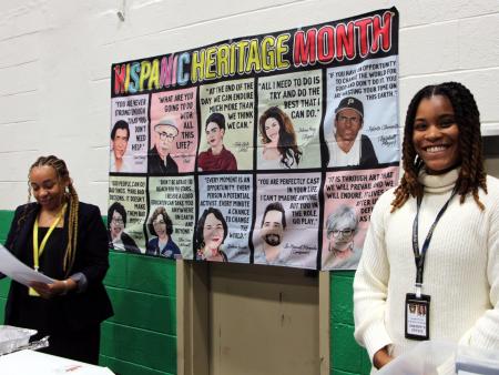 two civilian employees standing near a banner highlighting achievements of famous Hispanic people