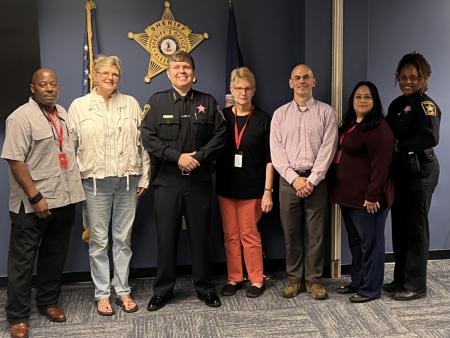 Seven people, including the Sheriff and a lieutenant in blue uniforms, standing in front of a U.S. flag, Virginia flag and a gold sheriff's star on a wall