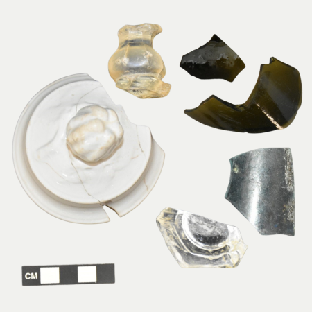 A collection of ceramic and glass artifacts recovered from a Lee Street Site privy. The artifacts include a white ceramic lid, clear table glass, and dark green bottle glass.