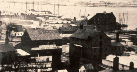 A black and white photo shows buildings along the waterfront with the masts of tall ships in the background. The Jamieson Bakery building is a three-story building with loft, circled in the foreground.