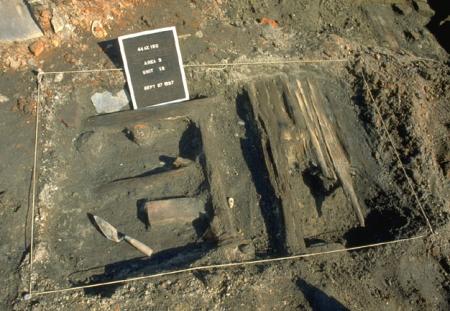 Photo of a rectangular Lee Street excavation unit with wooden floorboards exposed. String lines the outside of the unit, and a signboard and trowel label the unit and show which way is north.th.