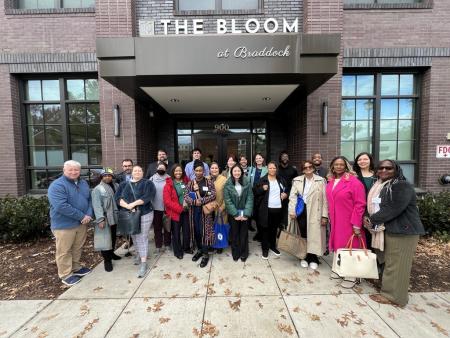 A group of people of differing ages, races, and genders in front of the entrance to a brown brick building. The sign over the entrance way reads "The Bloom at Braddock"