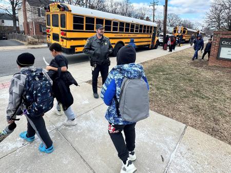 Deputy in a blue uniform watching as elementary students during dismissal. An orange school bus is in the background.