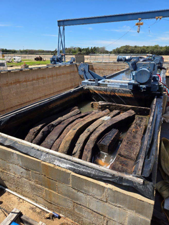 Wooden ship timbers sit partially submerged in a plastic-lined concrete tank. 