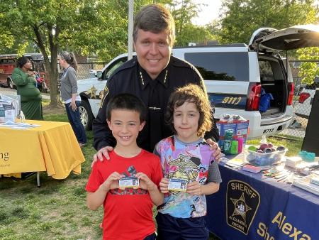 Sheriff in blue uniform with two kids who are holding up photo identification cards and smiling