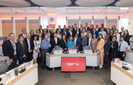 A group of people stand in front of the Bloomberg logo, smiling at the camera and dressed in business attire.
