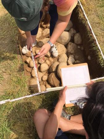 Two archaeologists are shown working in an excavation unit with a cobbled floor. One archaeologist uses a folding rule to measure where each cobble is located. The other archaeologist draws a map of the cobbles on a clipboard.