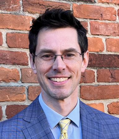 A man in business attire and glasses in front of a brick wall.