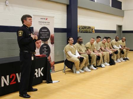 Sheriff in blue uniform praising nine inmates who are seated and wearing tan jumpuits