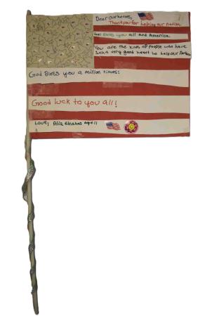A poster in the shape of an American flag on a stick reads, “Dear our heroes, Thank you for helping our nation. God Bless you all and America. You are the kinds of people who have such a very good heart to help our nation. God Bless you a million times! Good luck to you all! Love, Alia Elnahas age 11.”