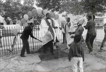 A Black minister in a white robe is caught in mid-air as he jumps on a symbolic grave marking the end of apartheid. Others dance and clap.