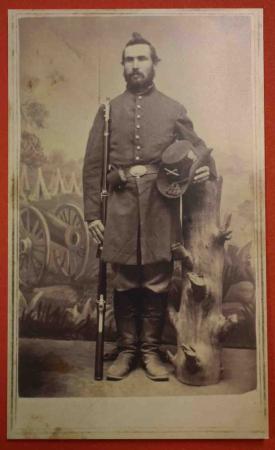 Private Francis E. Brainerd holds his rifle and his hat, leaning on a tree stump with a painted scene of a Civil War encampment behind him in this studio photograph.