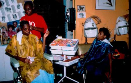 In this 1985 color photograph of a Black beauty shop, a hairdresser works on a patron while another looks on.