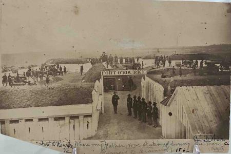 Men are lined up holding their rifles under the Fort O’Rourke sign. Other soldiers are standing in various places on the earthworks.