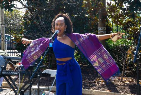 woman in blue jumpsuit with purple decorative shawl arms outstretched in front of a microphone