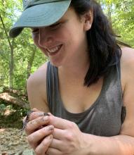 Coline smiles as she holds a very small brown-striped snake in her hands. The snake is crawling over her fingers and is holding its head out to the side, as if to sniff the air. Coline is in a forest with green leaves in the background, and is wearing a teal hat and a grey tank top, with her dark brown hair pulled back in a ponytail.