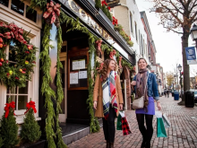 two women walking on brick sidewalk holding bags, standing in front of storefront decorated with garland, bows and a wreath